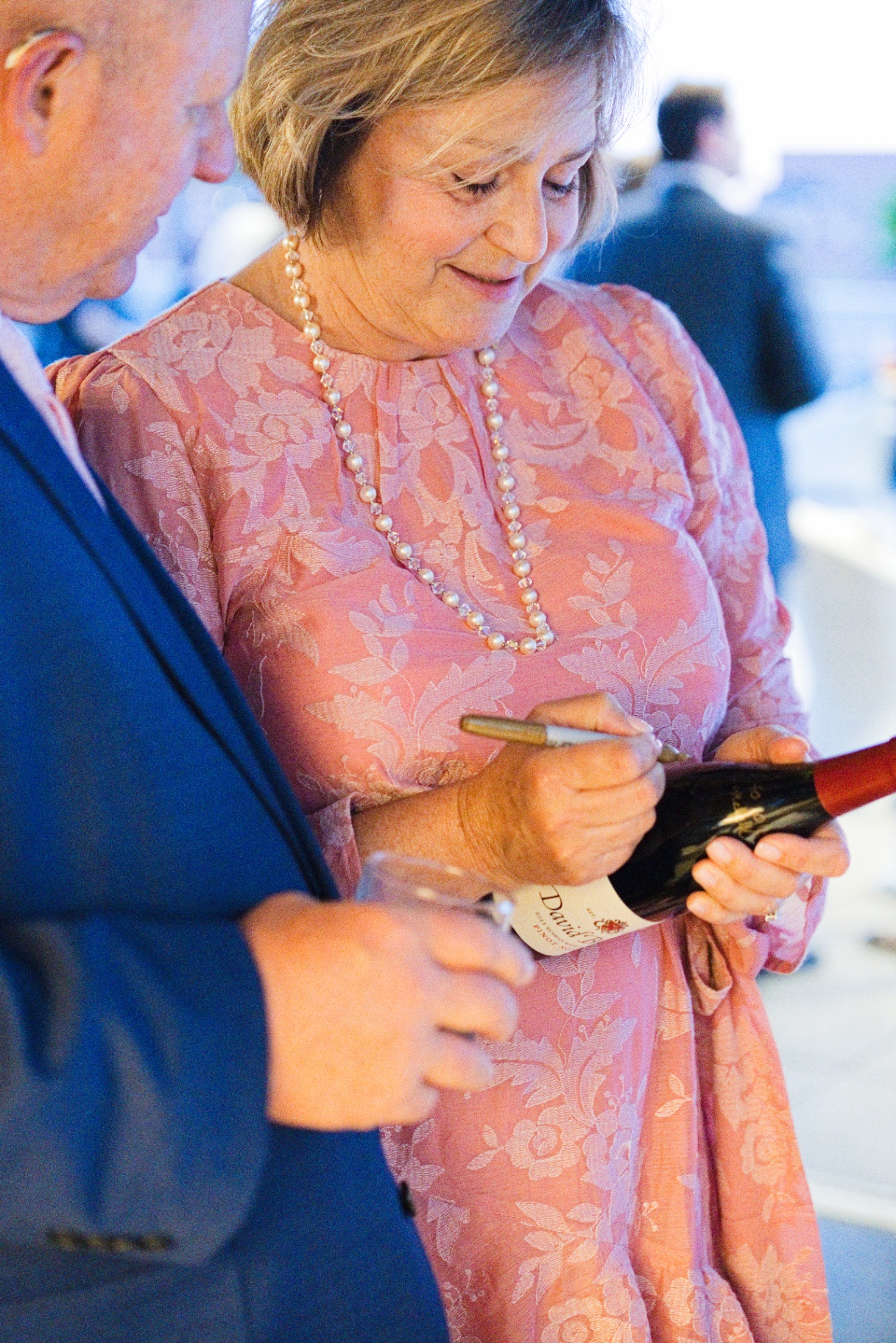 An elder wedding guest in a pink dress signing a David Bruce wine bottle with a gold pen